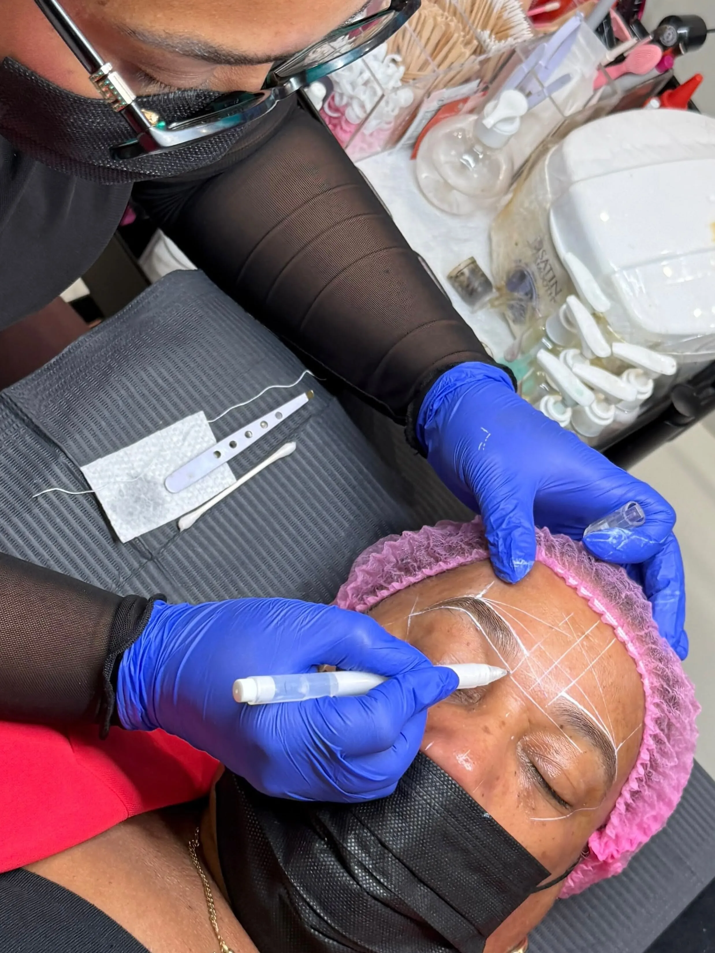Kathy mapping a client’s brow with a white pencil. The client wears a pink cap and a black mask, eyes closed.