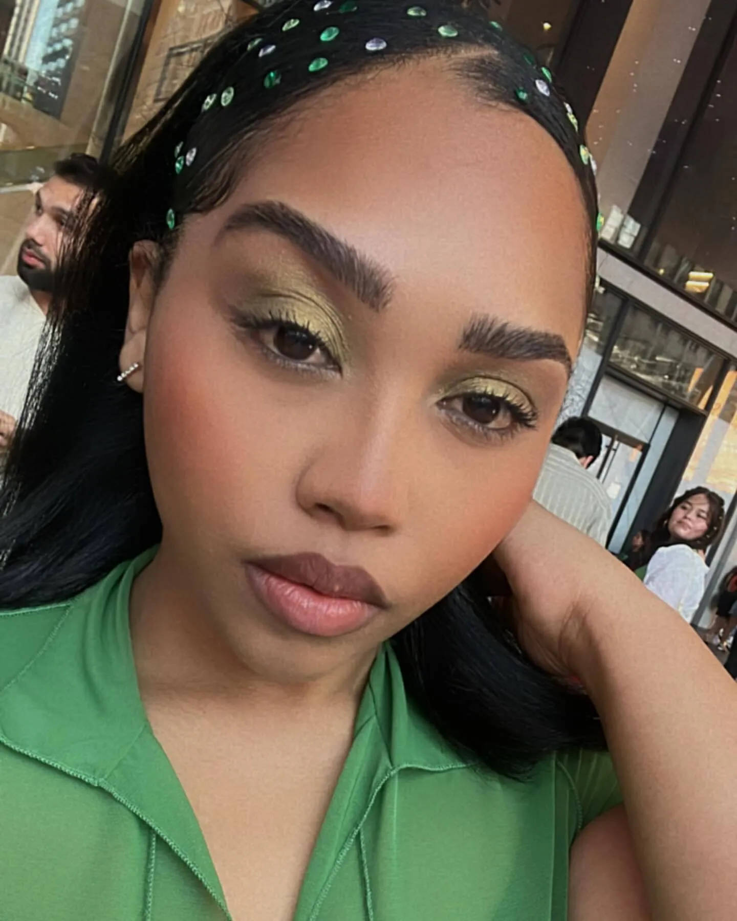 Close portrait in a green shirt, with gold-green eye makeup and defined brows.